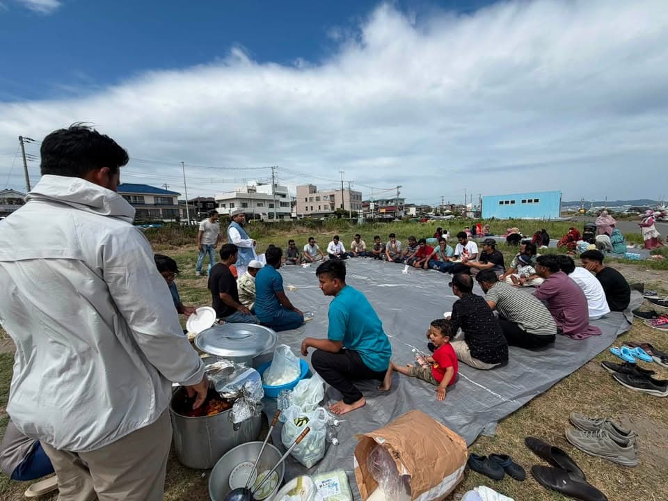 A large group of men and children sit together on tarps spread on grass near the coast, sharing a meal, with cooking pots in the foreground and shoes lined up at the edge of the tarps.