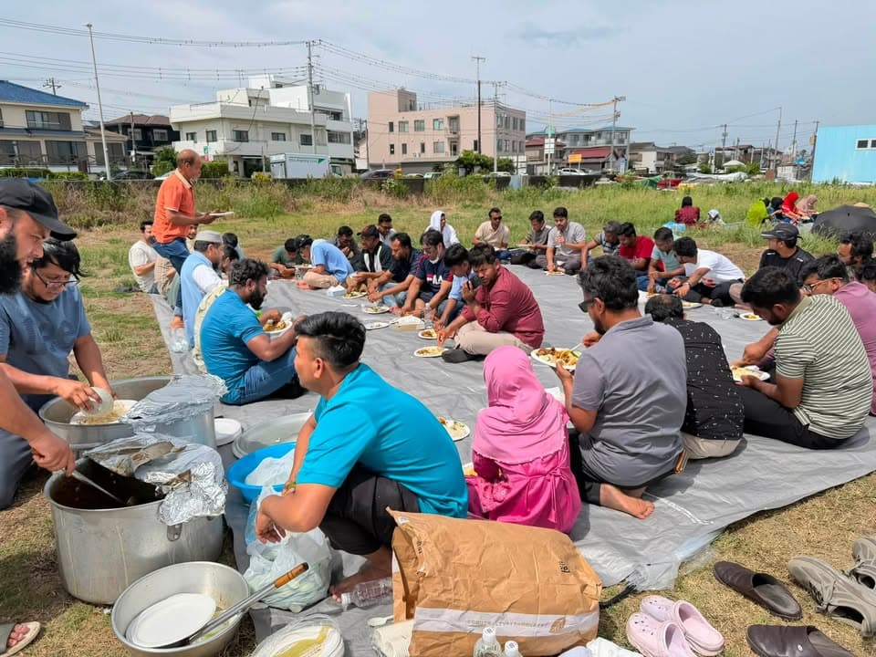 A large group of men and women sit on tarps spread over grass, eating from plates while food is served from big pots nearby, with residential buildings visible in the background.