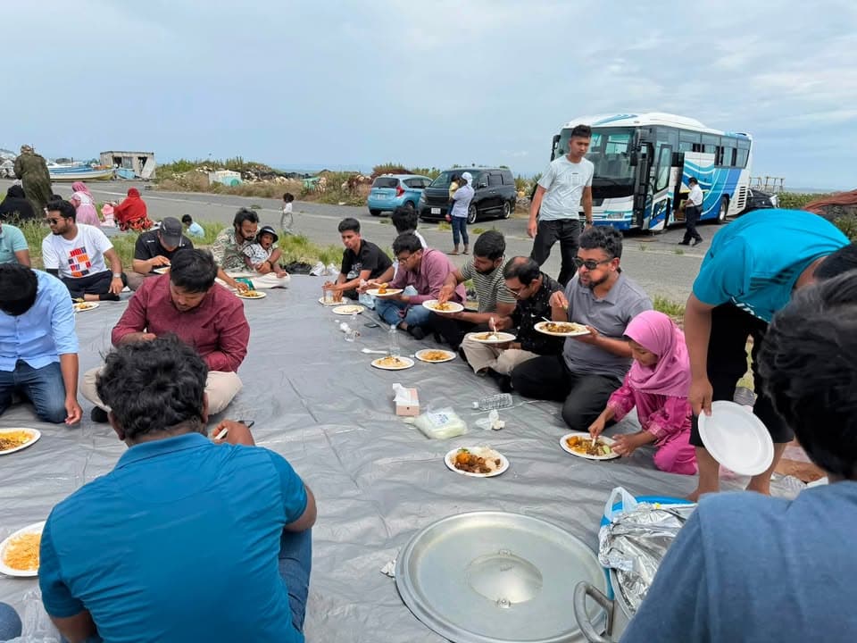 A group of men, women, and children sit on a large tarp spread on a coastal roadside, eating a meal together from paper plates, with a tour bus and cars parked behind them and the sea visible in the distance.