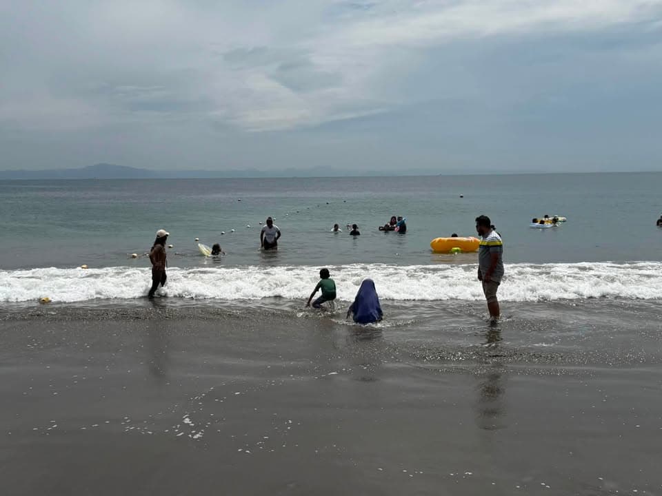 Families, including women in hijab and children, wade and play in the ocean waves at a sandy beach under a cloudy sky, with some using an inflatable float.