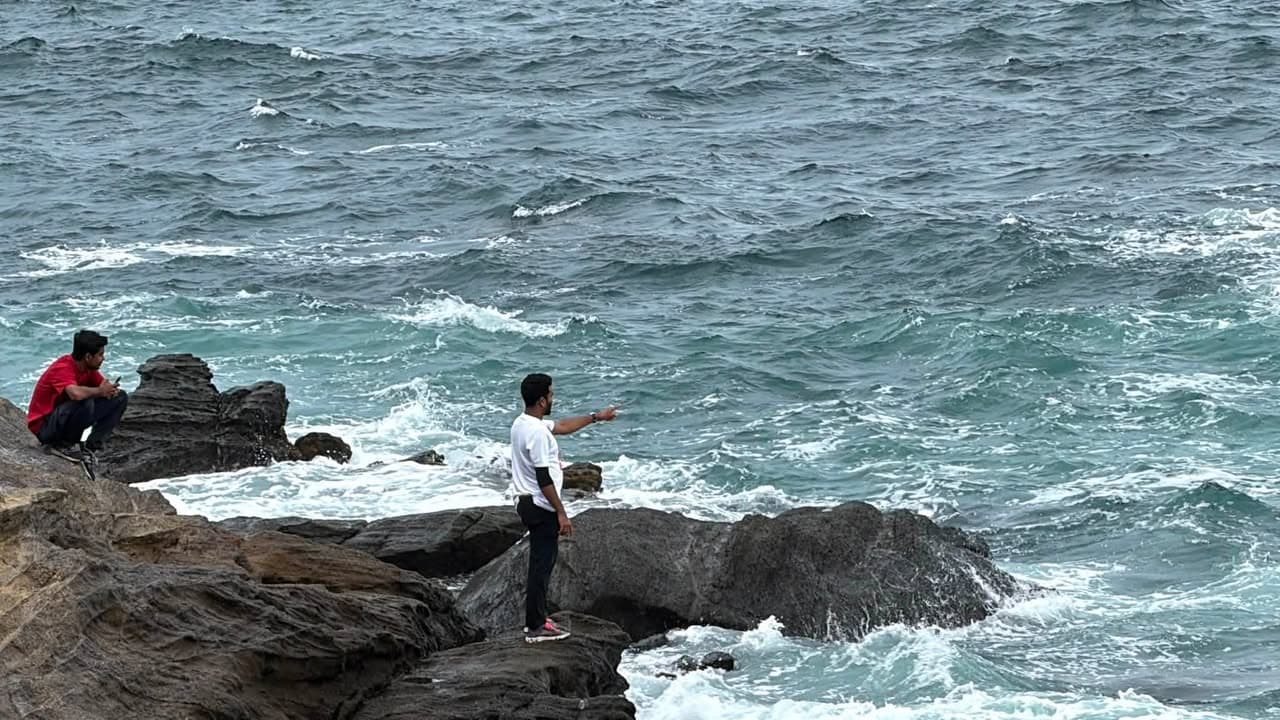 Two men stand on rocky coastal cliffs overlooking the ocean, one standing and pointing toward the sea while another sits nearby, with waves crashing against the rocks below.