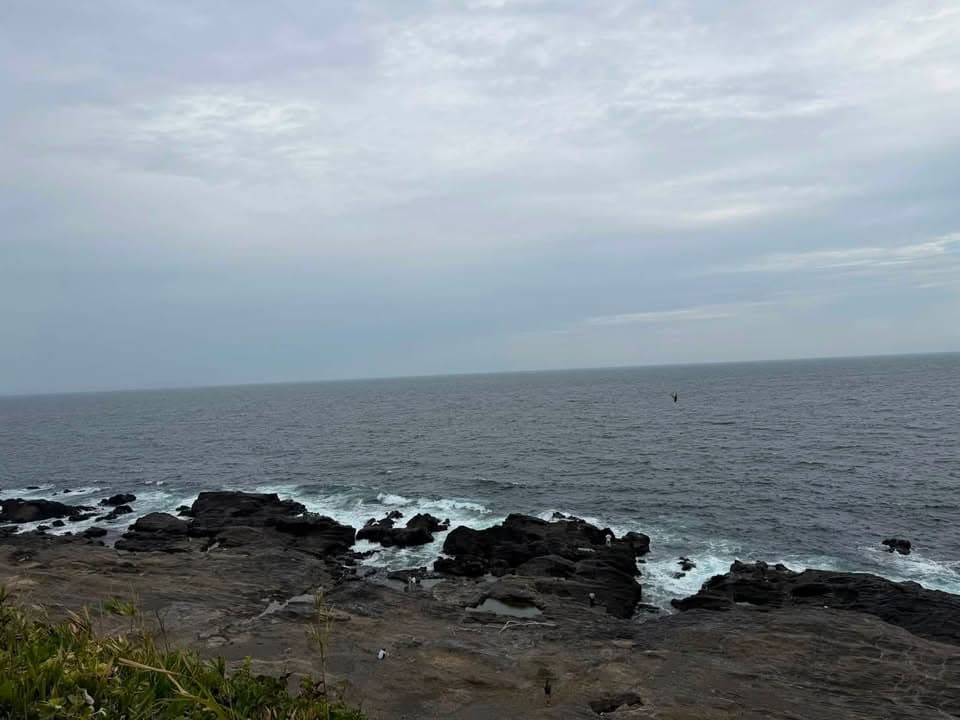 A rocky coastline with dark tide-worn rocks and crashing waves under a cloudy sky, with the open sea stretching to the horizon.