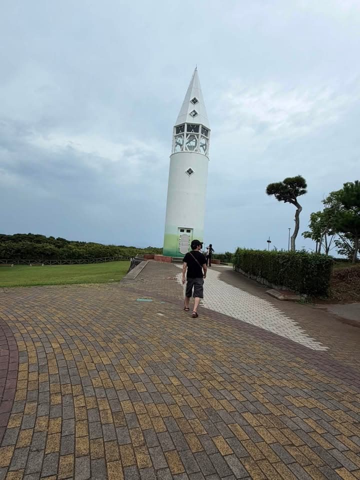 A person walks along a paved path toward a tall lighthouse with a conical roof, set on a grassy area near the coast under a cloudy sky.