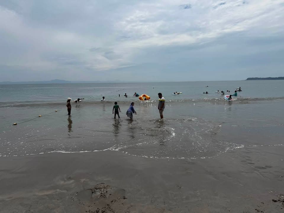 Adults and children wade and swim in shallow sea water at a beach under a cloudy sky, with some using inflatable floats further out.