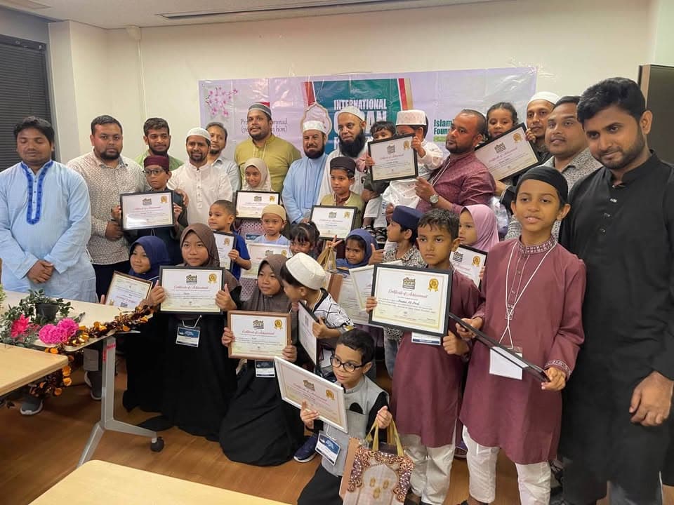 A group photo of children holding certificates of achievement alongside adult organizers and religious leaders, standing in front of an "International Qur'an" event banner in a community center room.