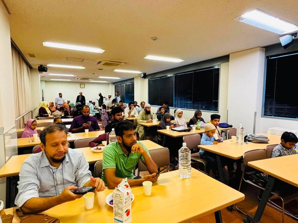 Men, women, and children sit at rows of tables in a community center hall, with drinks and snacks on the tables as some watch a program at the front of the room.