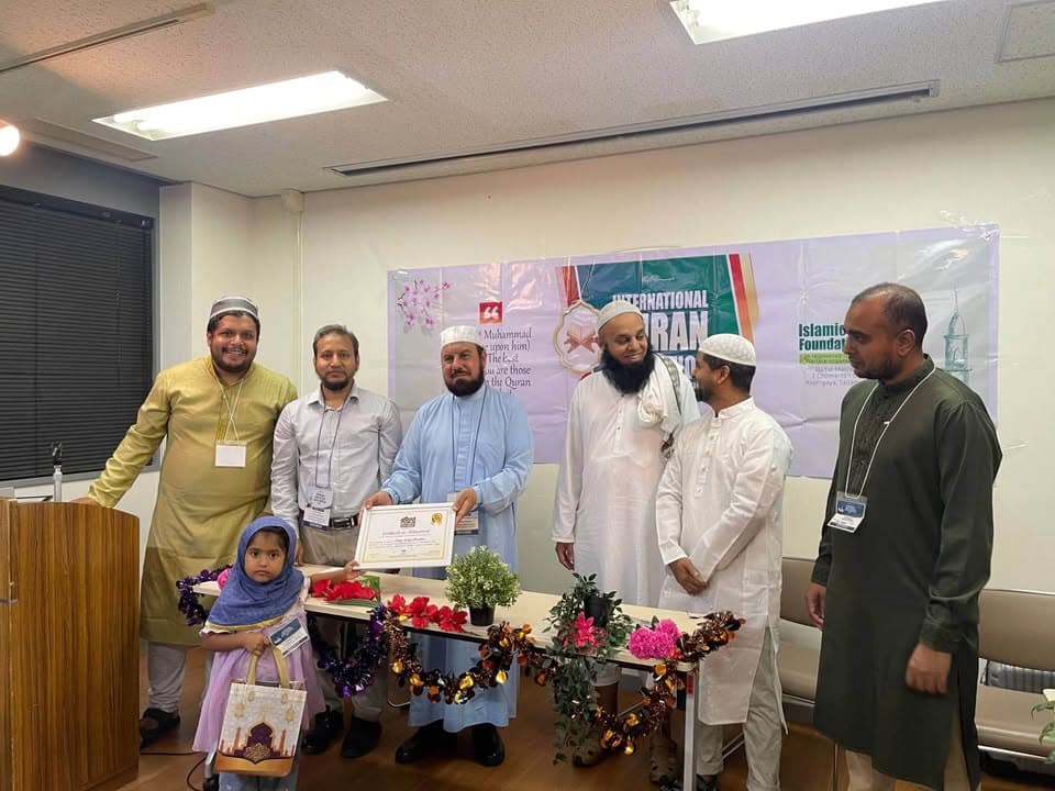 A group of men in traditional Islamic dress stand together in a conference room holding a certificate, in front of a banner reading "International Qur'an Competition" and "Islamic Foundation," with a table decorated with flowers, and a young girl in a headscarf standing beside them.