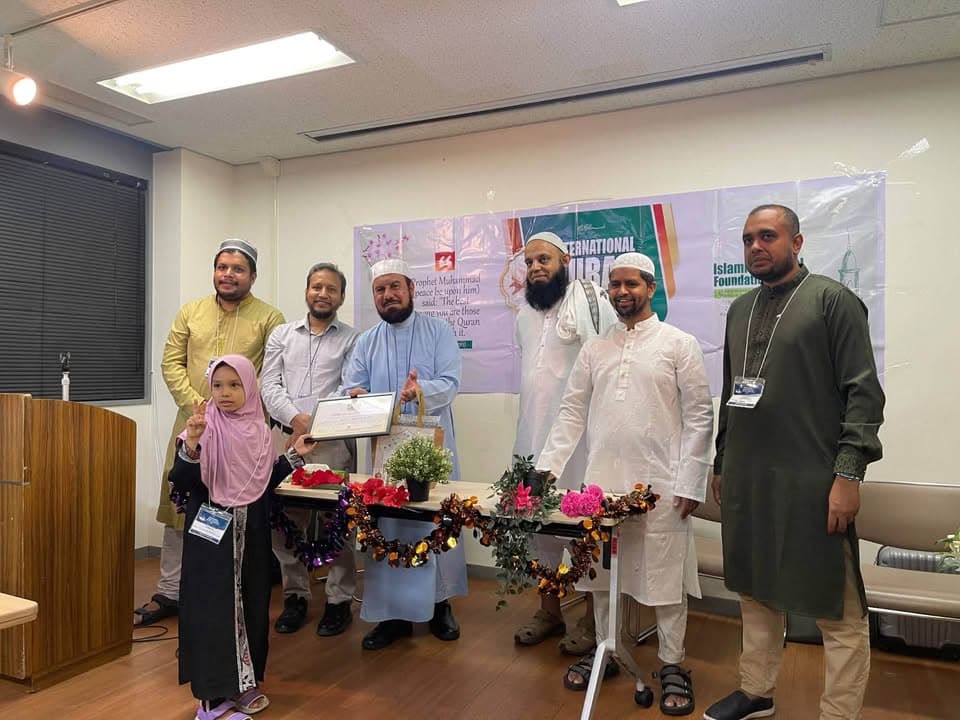 A group of men in traditional Islamic attire, a young girl in a pink hijab, and a man holding a certificate and gift bag stand together in front of an "International Qur'an" competition banner, with a decorated table of flowers before them.