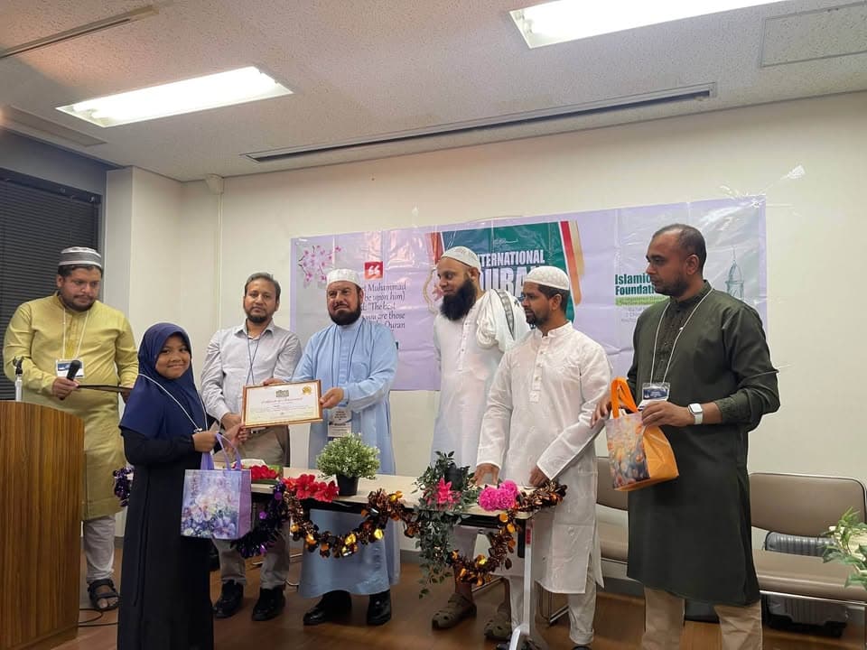 A young girl in a hijab holds a certificate presented to her by several men in traditional Islamic clothing, standing in front of an "International Qur'an" banner decorated with flowers on a table.