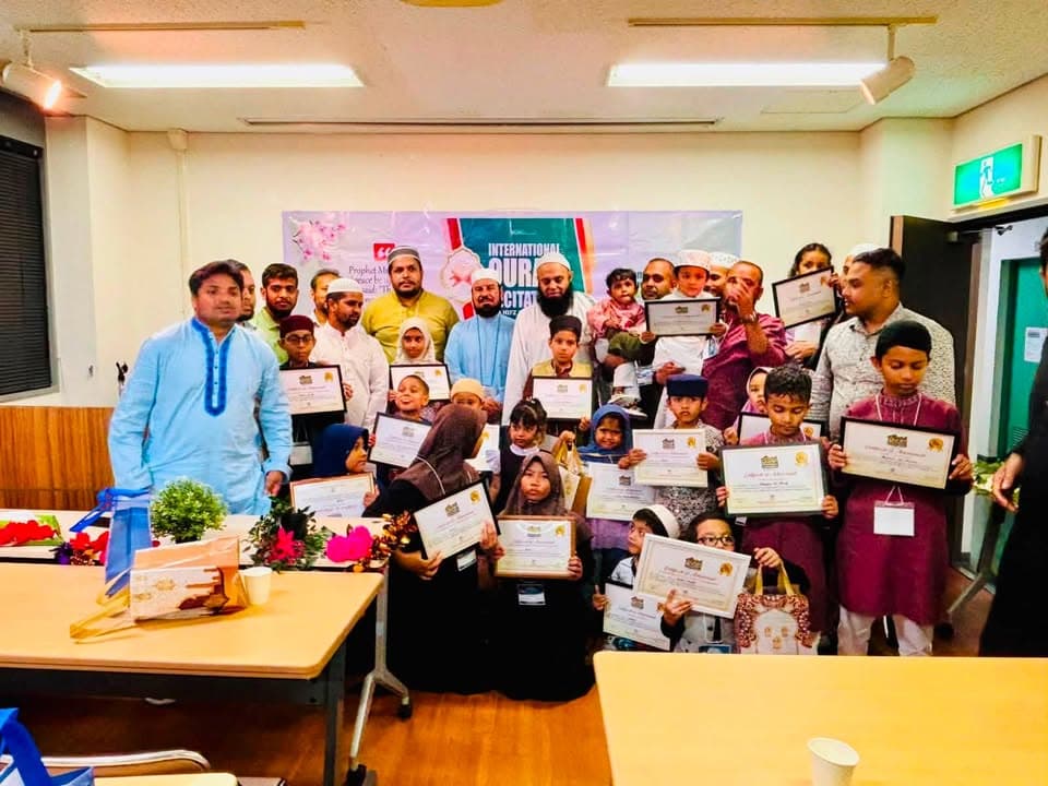 A large group of children and adults pose together indoors holding certificates, standing in front of a banner reading "International Qur'an Recitation and Hifz" competition.