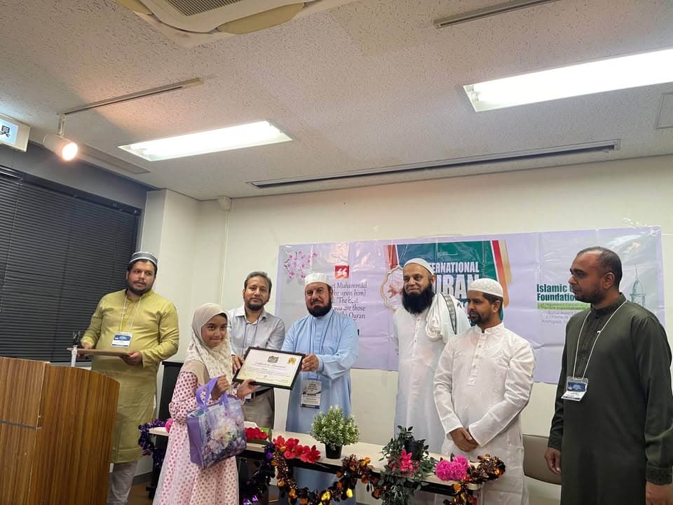 A young girl in a hijab holds a certificate presented to her by a group of men in traditional Islamic dress, standing in front of a banner reading "International Quran" and "Islamic Cultural Foundation," with flower decorations on a table in the foreground.