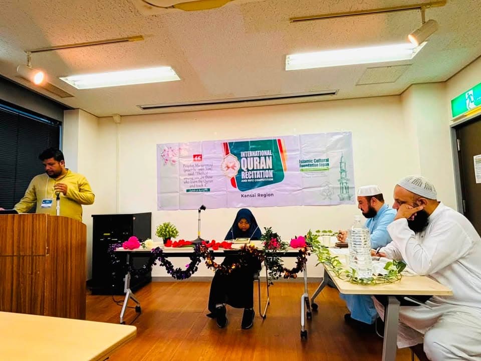 A young girl in a hijab sits at a decorated table to recite in front of a banner reading "International Quran Recitation and Hifz Competition, Kansai Region," while a man speaks at a podium on the left and judges sit at a table on the right.