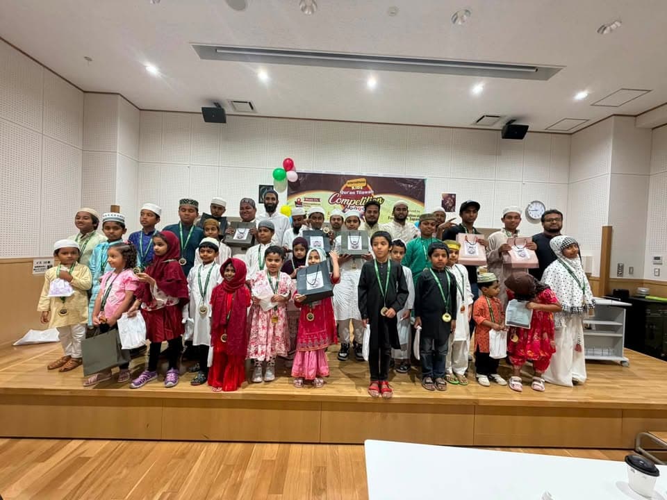 A large group of children and adult organizers stand together on a stage, many wearing medals and holding gift bags and boxes, in front of a "Kids Qur'an Tilawah Competition" banner with balloons.