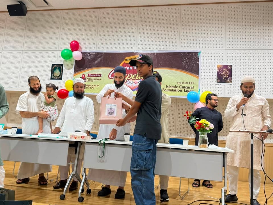 A young participant receives a gift bag from organizers on stage at a Qur'an competition event, with balloons and a banner reading "International Qur'an Competition" visible behind them, while other participants and a man speaking into a microphone stand nearby.