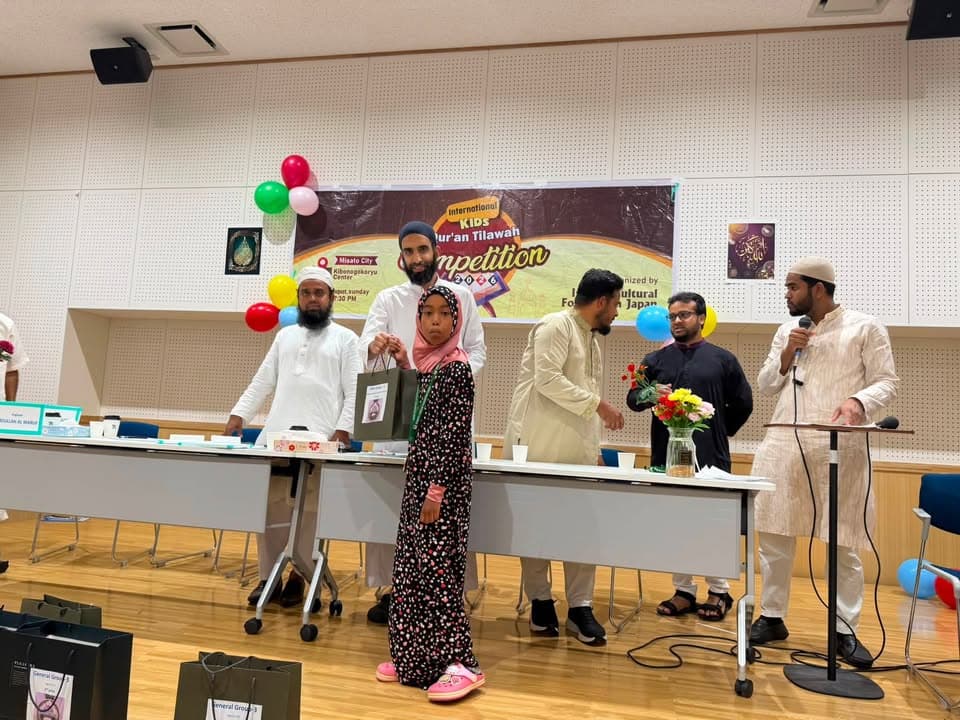 A young girl in a headscarf stands holding a gift bag on stage as a man beside her speaks, with judges seated at a table and a man announcing at a microphone, in front of a banner reading "International Kids Qur'an Tilawah Competition."