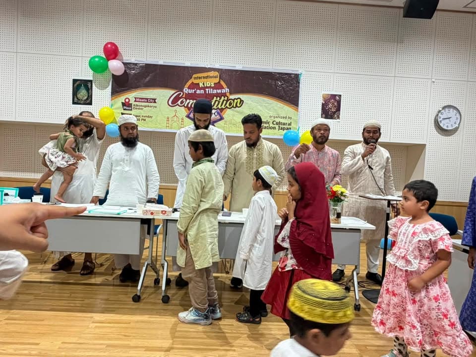 Children in traditional dress line up before a table of judges in a hall decorated with balloons and a banner reading "International Kids Qur'an Tilawah Competition," as men in white and beige robes look on.