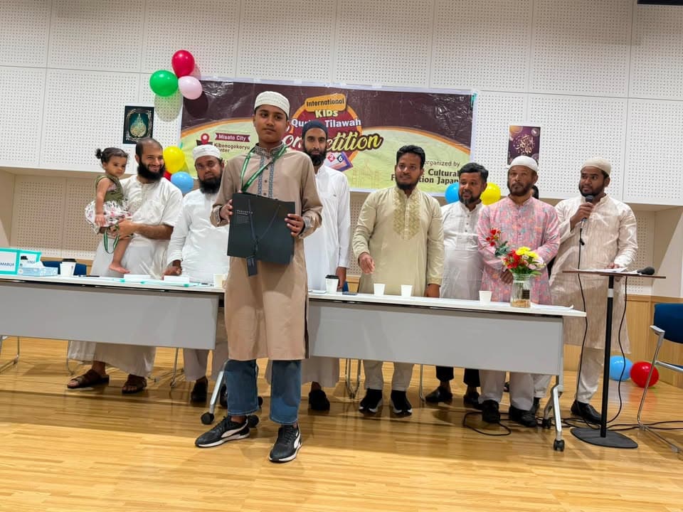 A boy wearing a cap and holding an award folder stands in front of a row of men, including judges and organizers, at an indoor event with a banner reading "International Kids Qur'an Tilawah Competition" and balloons in the background.