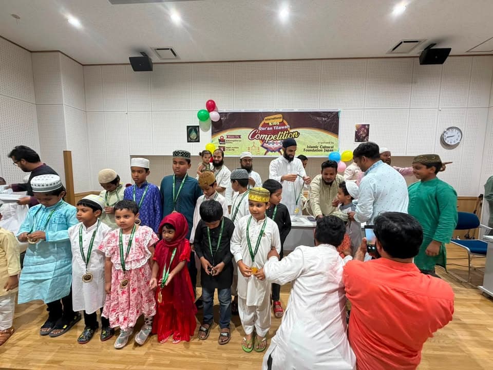 Children in traditional dress, many wearing medals, gather with adults on stage at an indoor hall in front of a banner reading "International Qur'an Tilawah Competition," decorated with balloons.