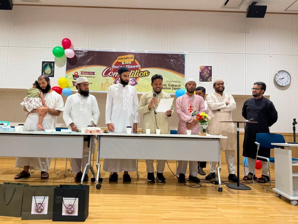 A group of men in traditional white and patterned attire stand behind a long table on a stage, one holding a microphone and reading from a paper, in front of a banner reading "International Kids Qur'an Tilawah Competition," with balloons decorating the wall and gift bags placed on the floor in front of the stage.