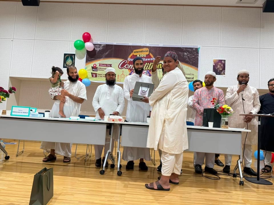 A young participant receives an award box while standing before a panel of men seated behind a long table decorated with balloons and a banner reading "International Kids Quran Tilawah Competition" at the Kibonogokoryo Centre.