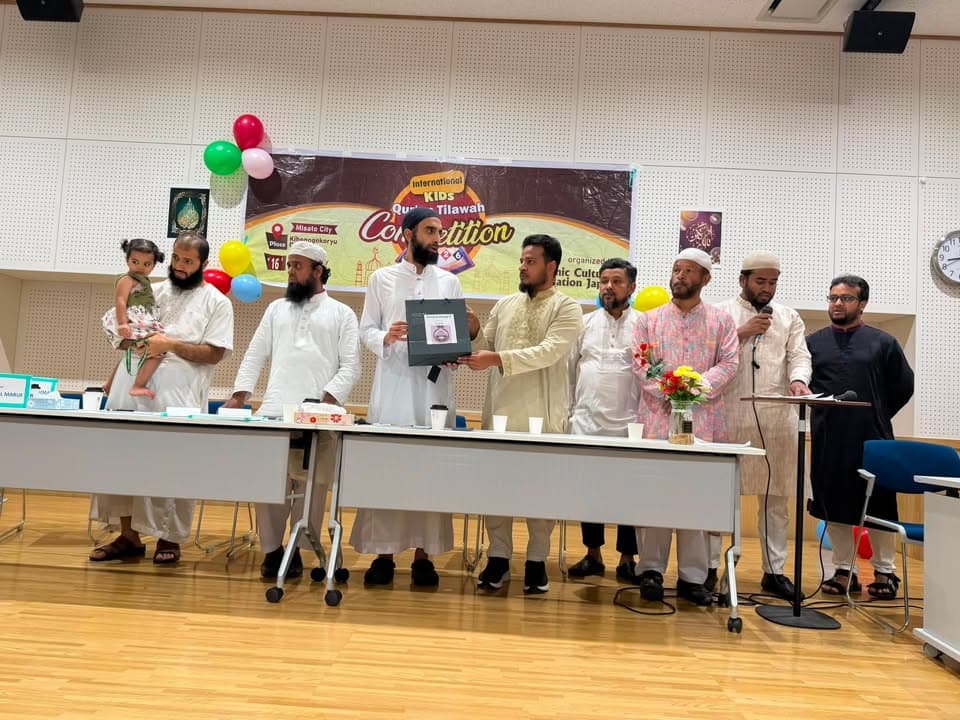 A group of men in traditional white and light-colored clothing, one holding a young child, stand behind a table presenting an award box, in front of a banner reading "International Kids Quran Tilawah Competition" decorated with colorful balloons.