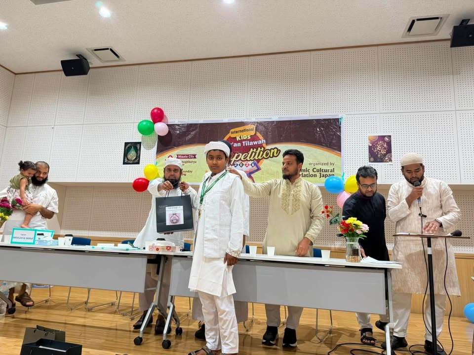 In a decorated hall with colorful balloons and a banner reading "International Kids Qur'an Tilawah Competition," a boy in white traditional dress stands at a long table receiving an award as several men in white and black robes stand beside him, one holding a microphone.