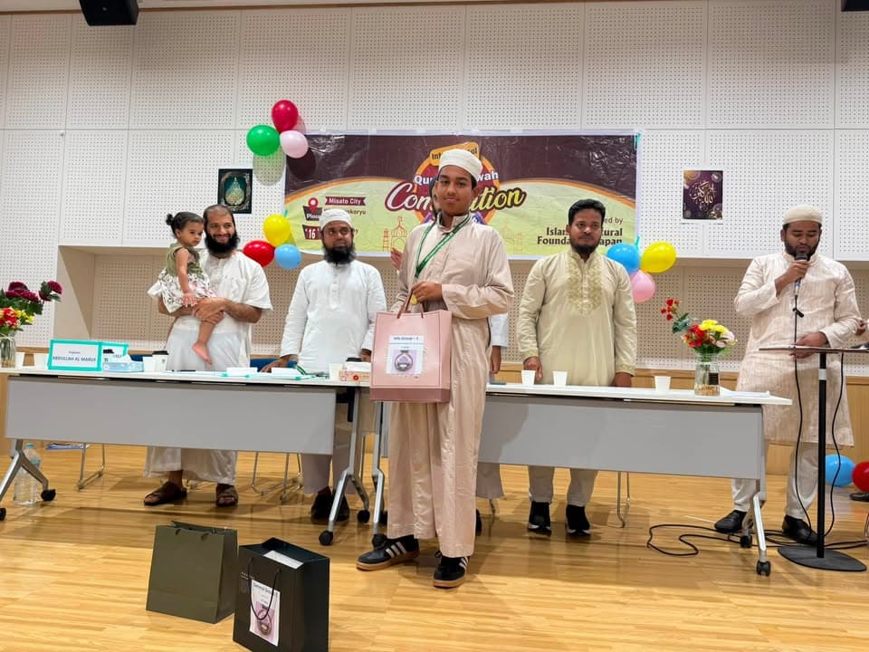A boy in a light-colored thobe and cap stands at the front of a hall holding a gift bag, while several men in traditional white attire stand behind a long table with balloons and a competition banner reading "International Qur'an Recitation and Hifz Competition" in the background; one man at a podium speaks into a microphone.