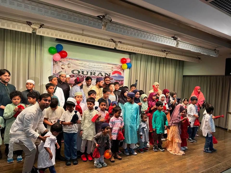 A large group of children in traditional Islamic dress, along with several adult men, pose together on a stage decorated with colorful balloons and a banner reading "International Quran Recitation and Hifz Competition," with many children holding small gift bags.