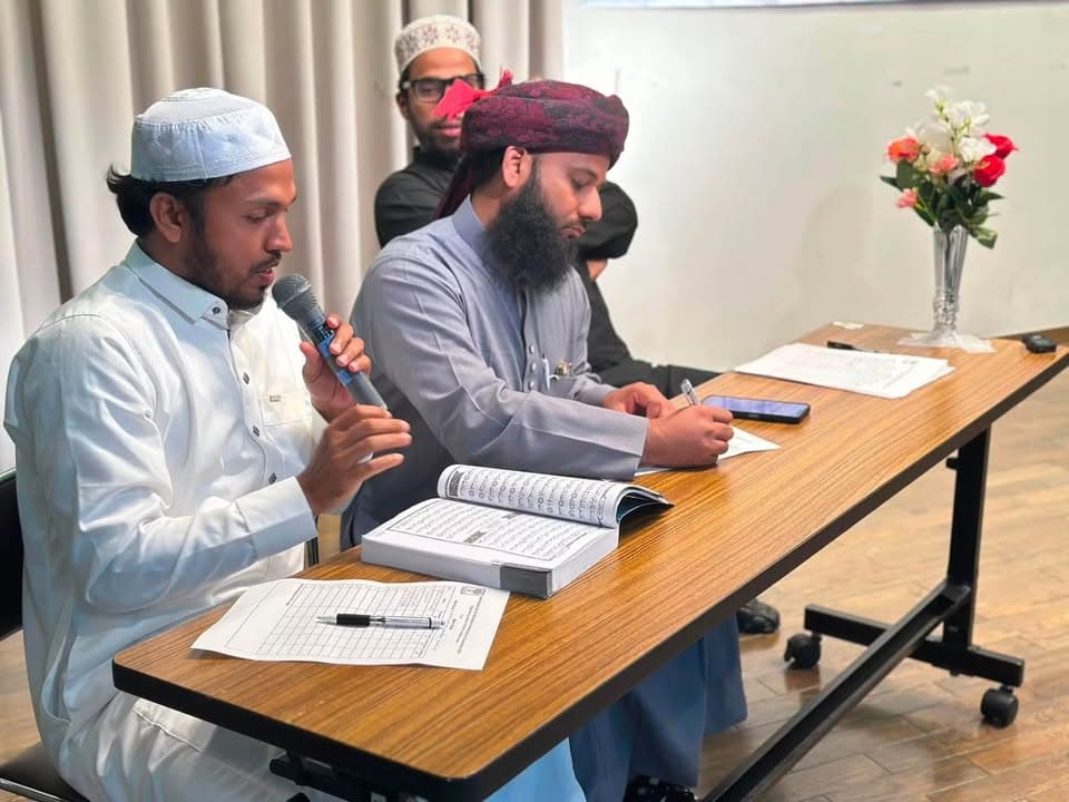 A man in a white cap speaks into a microphone while reading from an open Qur'an at a long table, as another man beside him wearing a maroon turban writes on paper, with a third man standing behind them and a vase of flowers on the table.