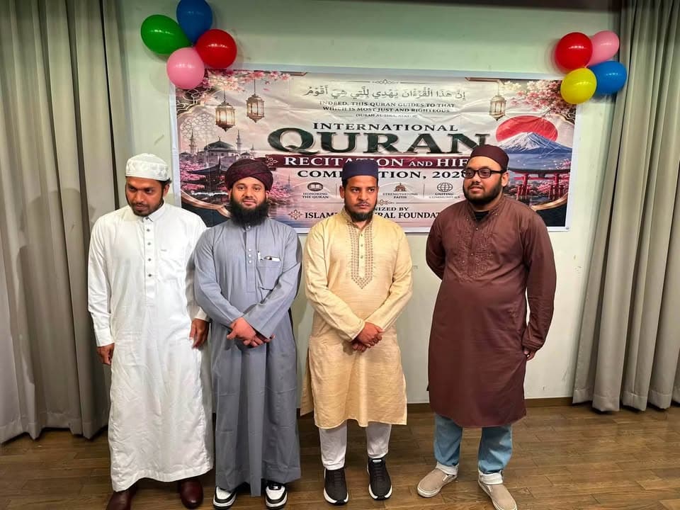 Four men in traditional Islamic attire, including caps and a turban, stand in a row in front of a decorated banner reading "International Quran Recitation and Hifz Competition 2026," flanked by colorful balloons and green curtains.