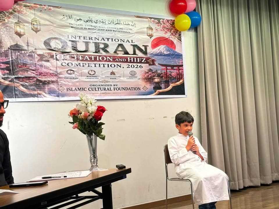 A young boy in a white thobe sits on a chair holding a microphone and reciting, in front of a banner for the International Quran Recitation and Hifz Competition 2026, decorated with balloons and a flower vase on a judges' table.