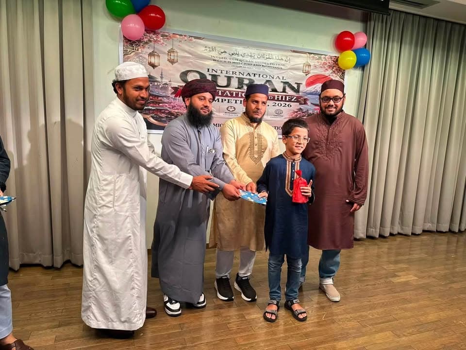 Four men in traditional robes and caps stand on stage with a young boy wearing glasses, presenting him with a gift in front of a backdrop reading "International Quran Recitation and Hifz Competition 2026," decorated with colorful balloons.