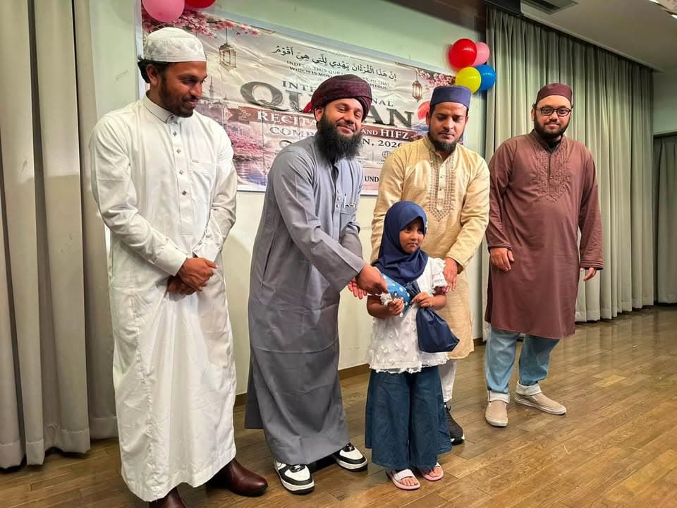 A young girl in a hijab receives a prize from a bearded man in a gray robe, while three other men in traditional Islamic attire stand beside her on a stage decorated with balloons and a Qur'an competition banner.