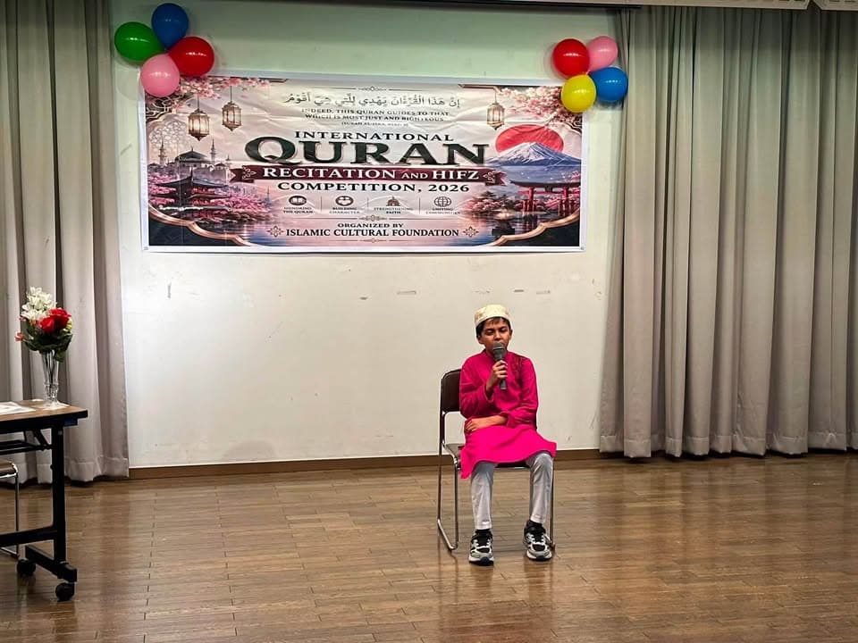 A boy wearing a pink kurta and white cap sits on a chair on a stage, reciting into a microphone in front of a banner reading "International Quran Recitation and Hifz Competition, 2026" decorated with colorful balloons.