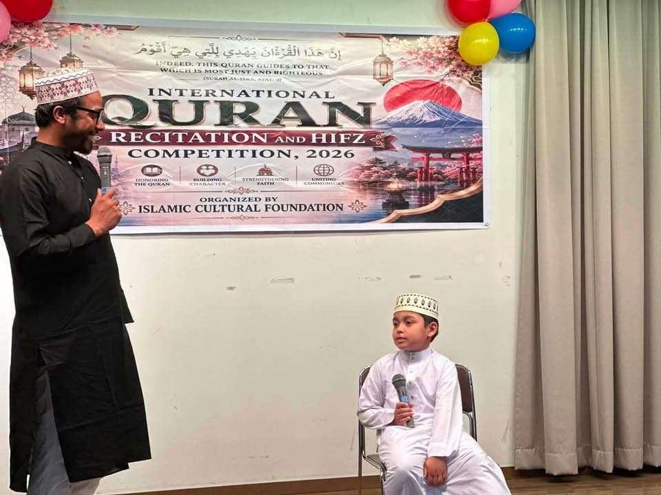 A man in a black kurta and cap holds a microphone and speaks while standing beside a seated young boy in white traditional dress and a cap holding a microphone, in front of a large banner reading "International Quran Recitation and Hifz Competition, 2026, Organized by Islamic Cultural Foundation," with balloons decorating the wall.