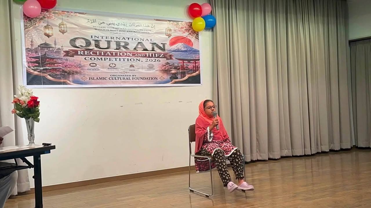 A young girl in a pink headscarf sits on a chair on stage, holding a microphone as she recites, in front of a large banner reading "International Quran Recitation and Hifz Competition, 2026" decorated with balloons.
