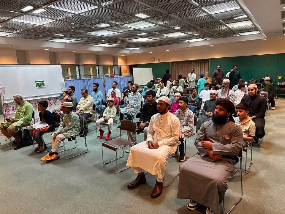 Men and boys in traditional attire, many wearing prayer caps, sit in rows of chairs facing forward in a large hall with a green wall and whiteboards, attentively watching an event.