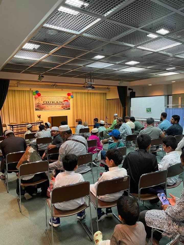 Men, boys, and children sit on rows of chairs facing a stage decorated with balloons and a banner reading "QURAN" at an indoor hall event.