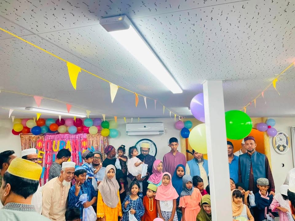 A large group of men, women, and children in traditional Islamic dress pose together in a room decorated with colorful balloons and bunting, with a "Qur'an Recitation" sign visible in the background.