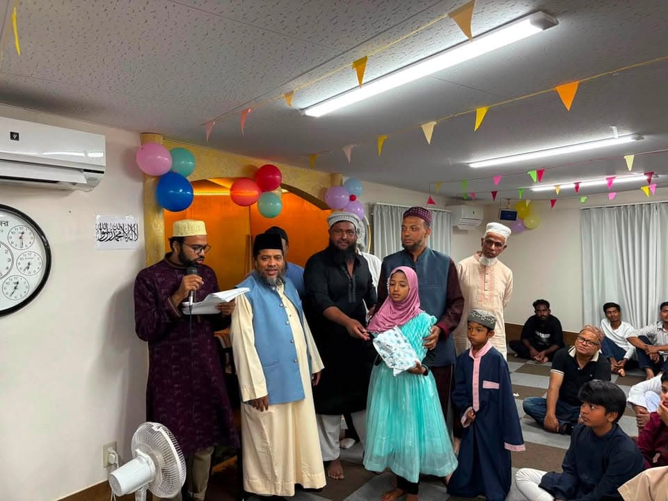 Inside a decorated prayer hall with colorful balloons and paper garlands, a man holding a microphone speaks while several men and children stand beside him, and a girl in a pink and teal dress is presented with a gift, as other attendees sit on the carpeted floor watching.