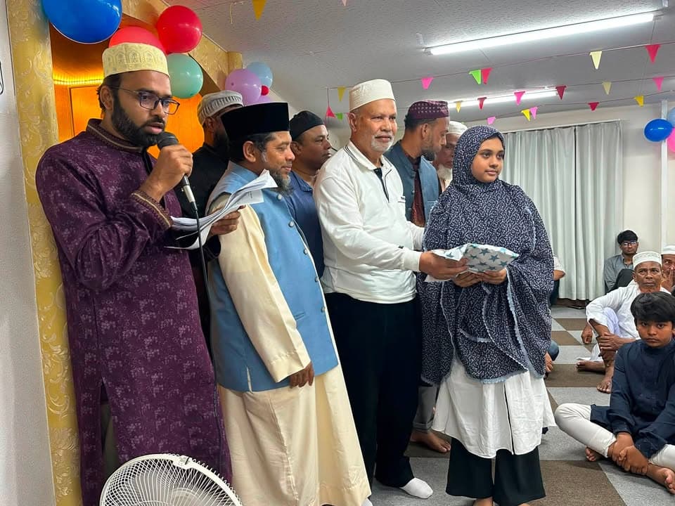 A man in a purple robe and cap speaks into a microphone while an elderly man in white presents a wrapped gift to a young girl in a headscarf, surrounded by other men in traditional attire in a room decorated with balloons and colorful bunting.