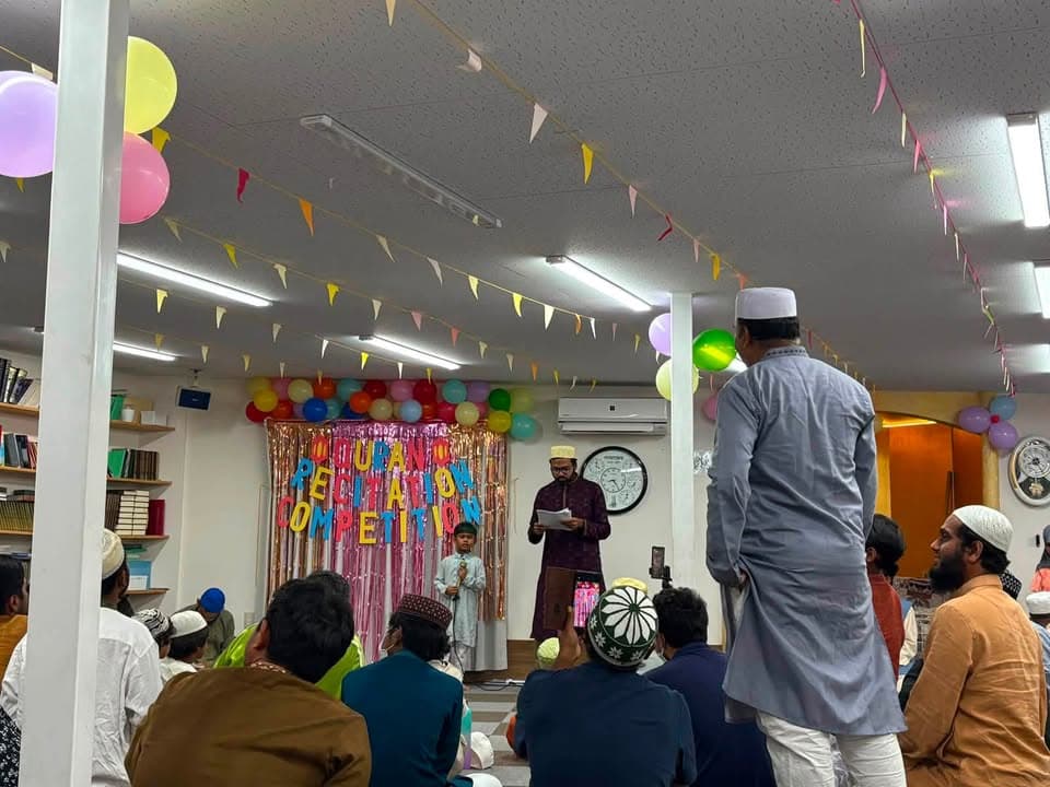 A boy stands at a microphone reciting the Qur'an on a stage decorated with balloons and a "Quran Recitation Competition" banner, while a man beside him reads from papers and an audience of men and boys sits on the floor watching.