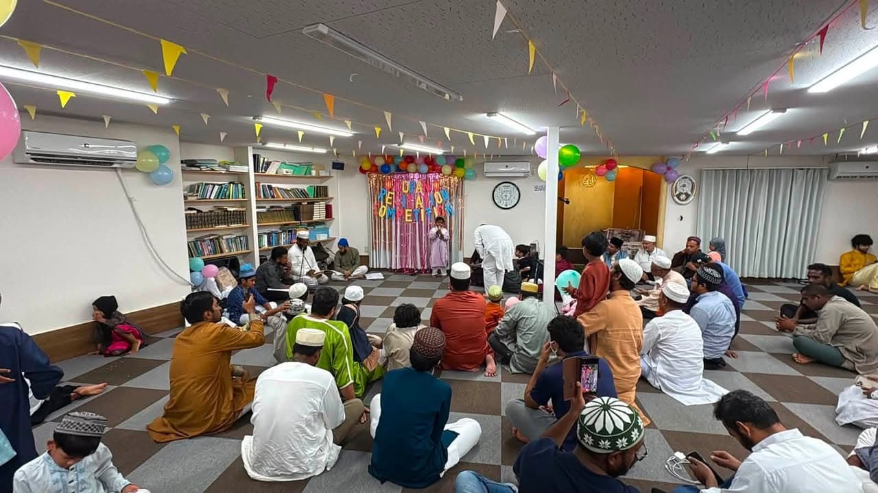Children and adults sit cross-legged on a checkered floor inside a decorated mosque hall, facing a small stage with balloons, banners, and a colorful backdrop reading "Quran Recitation Competition," while a child stands at a microphone reciting.