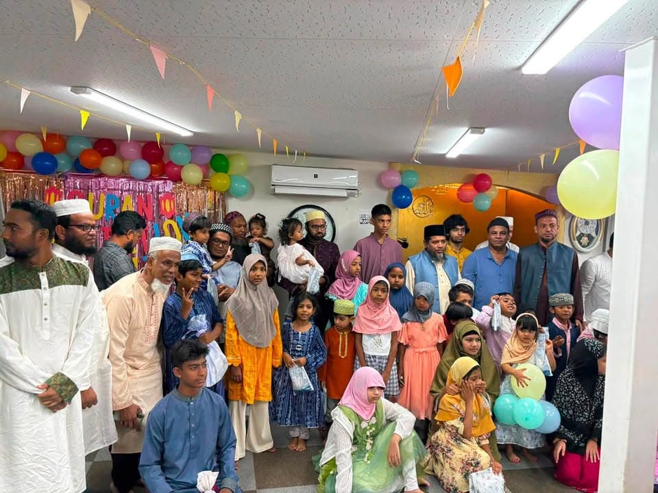 A large group of men, women, and children in traditional dress pose together for a group photo in a mosque hall decorated with colorful balloons, bunting flags, and a "Qur'an" banner.