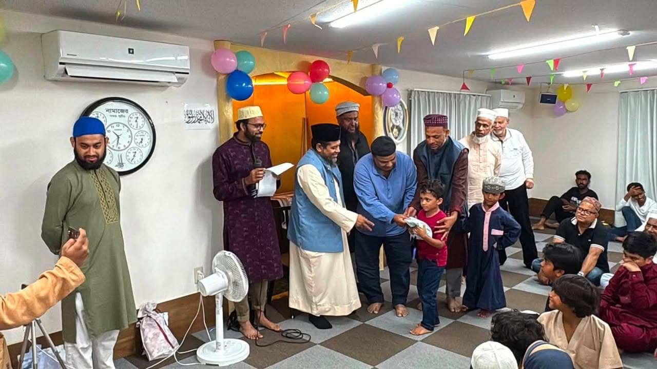 Inside a mosque decorated with colorful balloons and paper garlands, men in traditional dress present an envelope to a young boy while other participants and attendees look on, seated on a checkered carpet.