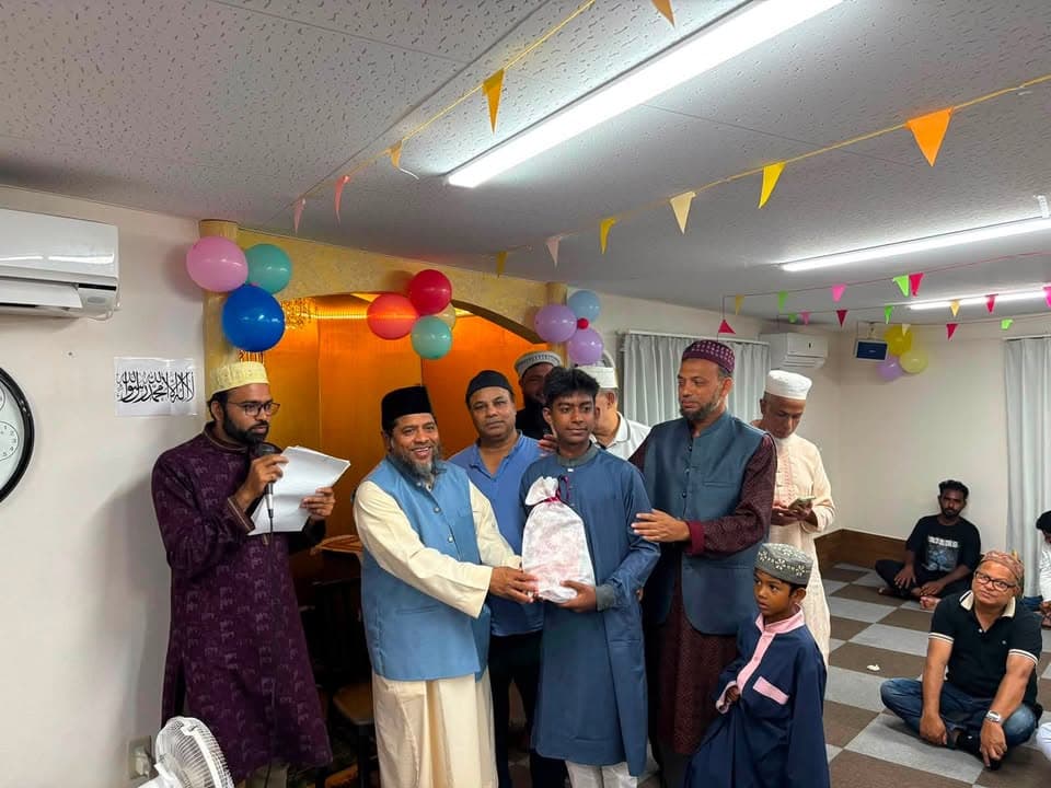 Inside a mosque hall decorated with colorful balloons and paper bunting, a group of men in traditional clothing present a wrapped gift to a young man, while others including a boy and seated attendees look on.