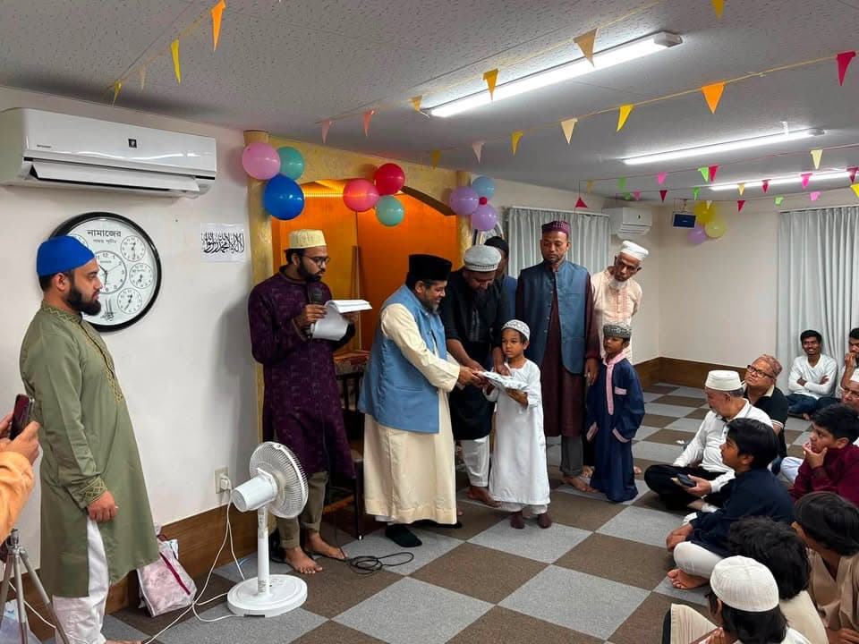Inside a room decorated with colorful paper bunting and balloons, a group of men and boys in traditional caps and robes stand as one man presents a certificate to a young boy, while others watch seated on the checkered floor.