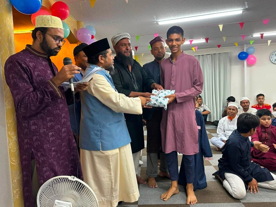 In a balloon-decorated mosque hall, men in traditional dress present a wrapped gift to a teenage boy while other men and children sit on the floor watching.