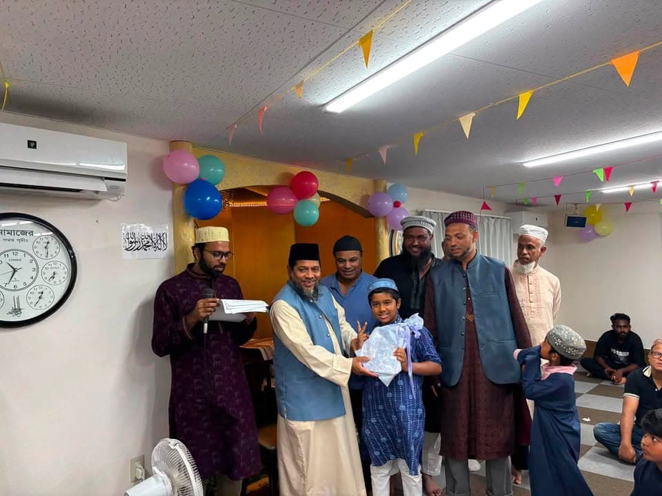 Inside a mosque hall decorated with colorful balloons and paper pennants, a group of men in traditional attire stand together as a boy holding a wrapped gift makes a peace sign, while a man beside him holds a microphone and papers.