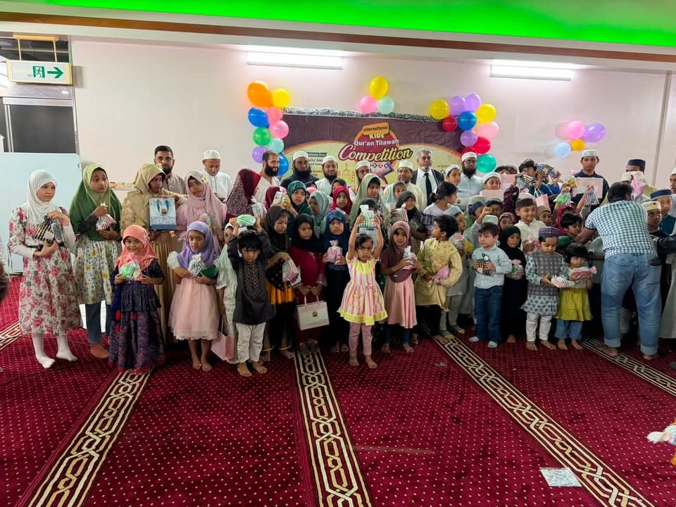 A large group of children and adult organizers pose together in a mosque prayer hall decorated with colorful balloons and a banner reading "International Kids Qur'an Tilawah Competition," with many children holding small gift bags.